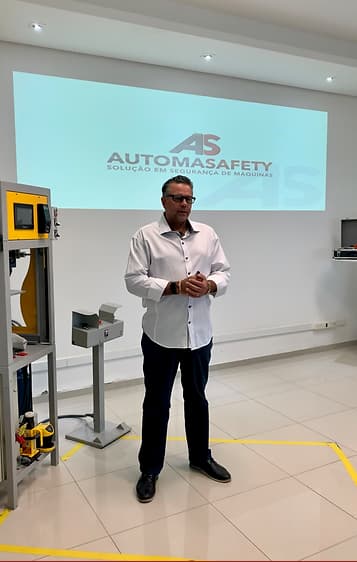Vladimir Kuse, Diretor da Automasafety em uma sala ao lado de uma máquina com pequenos três andares. Apenas no topo da máquina há uma tela semelhante a de um celular e um aparelho com vários botões ao lado. Nas duas outras áreas tem um espaço aberto. Vladimir usa sapato, calça e camisa social. Há um telão na sala escrito: A ésse Automasafety, solução em segurança de máquinas. 