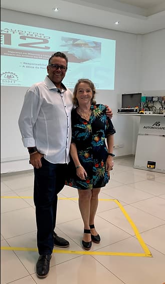 Vladimir Kuse, Diretor da empresa Automasafety ao lado de Luísa Tânia, Engenheira Civil e de Segurança do Trabalho. Eles possuem conhecimento sobre a Norma Regulamentadora Número doze e estão em uma sala de frente para um telão com informações escritas. A Engenheira tem cabelo curto e usa colar e blusa com flores ilustradas. O Diretor usa óculos e está com uma camisa de mangas compridas. Os dois estão sorrindo.